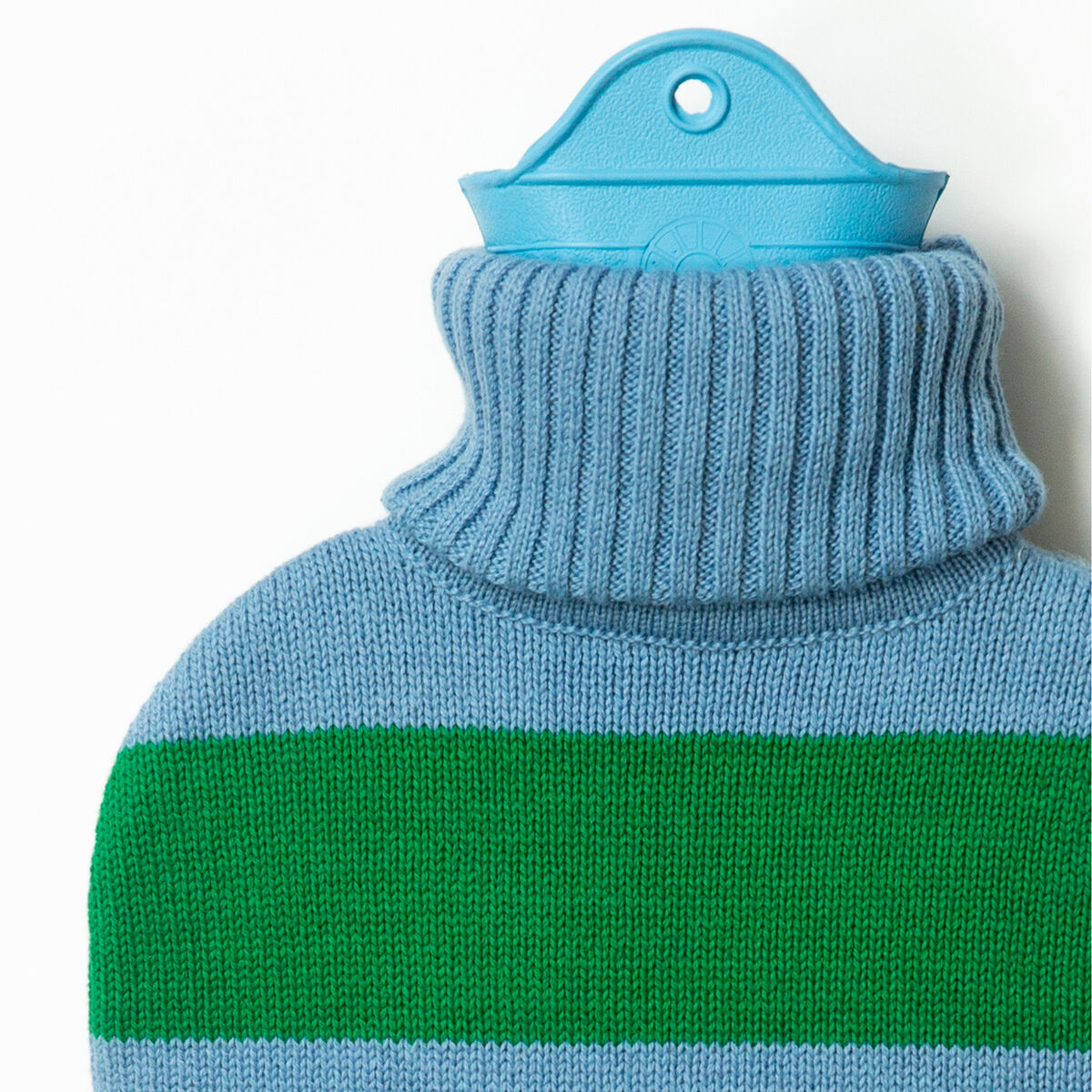 Detailaufnahme einer von SUITE702 hergestellten Wärmflasche mit gestricktem Bezug. Der Bezug ist blau mit einem breiten grünen Streifen und Rollkragen. Die Flasche darunter ist hellblau. Die Szene ist klar ausgeleuchtet, vor weißem Hintergrund.