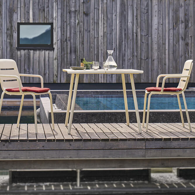 Ein moderner Gartentisch des Modells VAPIO, Größe 90x90 cm, steht auf einer Terrasse neben einem Pool. Der Tisch ist aus hellen Materialien gefertigt und wird von zwei passenden Stühlen mit roten Sitzkissen flankiert. Im Hintergrund ist eine Holzwand zu sehen. Ein stilvolles und minimalistisches Design.