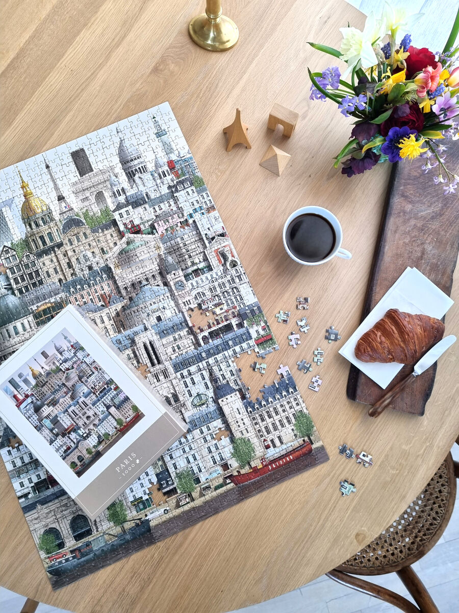 Das Bild zeigt ein 1000-teiliges Puzzle mit dem Motiv von Paris auf einem Holztisch. Neben dem Puzzle befinden sich einige verstreute Puzzleteile, eine Tasse Kaffee, ein Croissant auf einem Teller und ein Vasenstrauß mit bunten Blumen. Im Hintergrund sind goldene Dekorationen und ein paar Holzkunstwerke zu sehen.