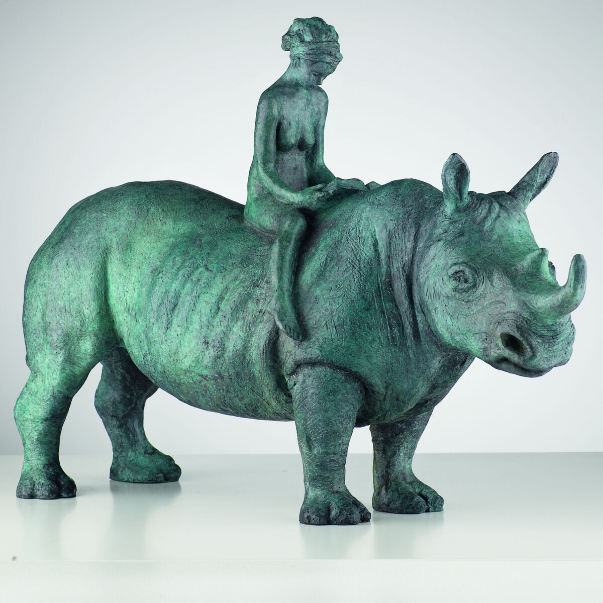 Patinierte Bronzeskulptur „The Maid and the Unicorn“ von Alfredo Llorens García: Ein kräftiges Nashorn mit detailreicher, rauer Haut steht seitlich auf weißem Grund. Auf seinem Rücken sitzt aufrecht eine schlanke nackte Frau mit hochgestecktem Haar; ihre Hände ruhen vor ihr auf dem Tier.