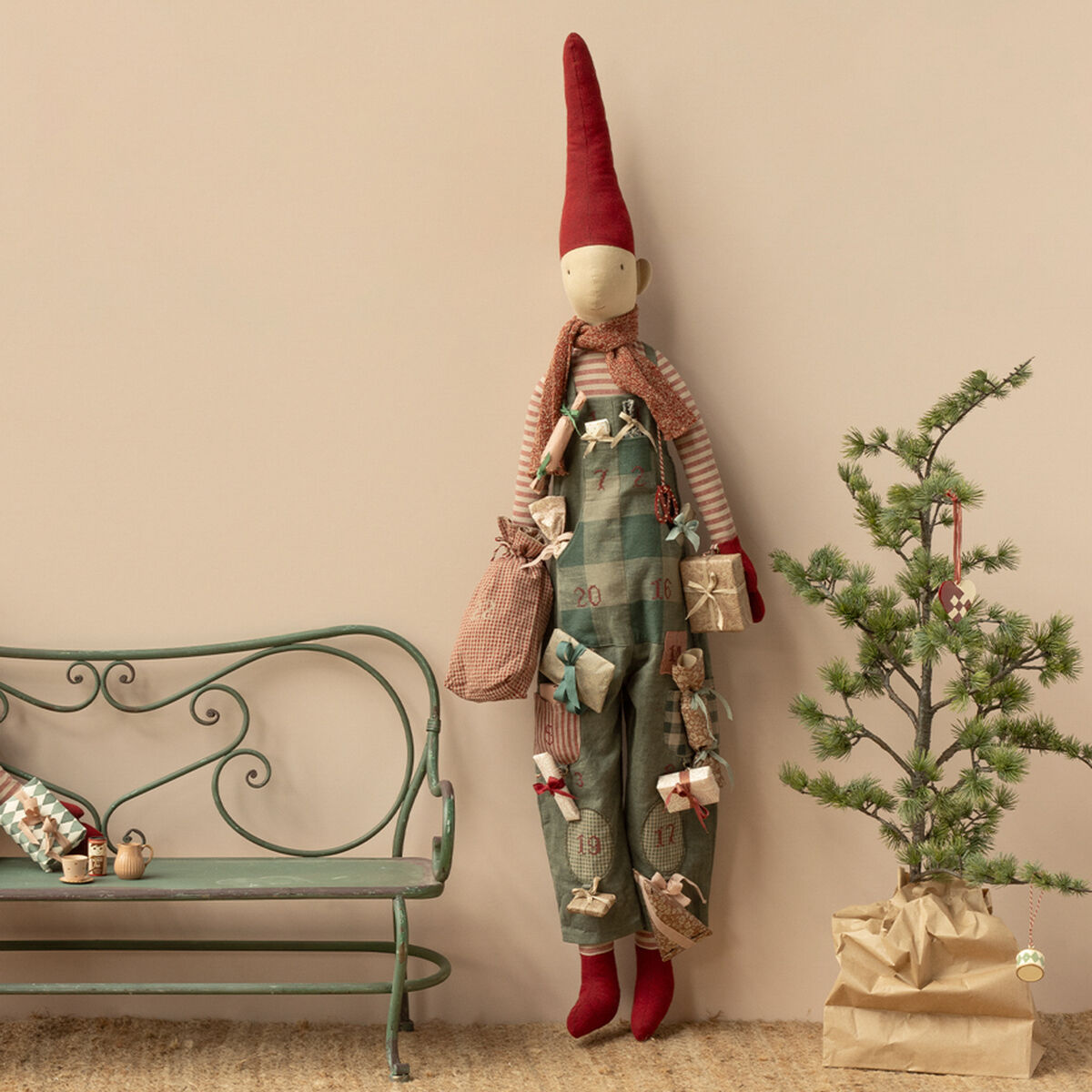 Zwei skandinavische Stoffpuppen im Pixy-Junge-Stil tragen Latzhosen mit roten Mützen und sind als Adventskalender dekoriert. Eine Puppe sitzt neben kleinen Geschenkpäckchen auf einer grünen Bank, die andere steht daneben. Im Hintergrund steht ein kleiner Weihnachtsbaum in einer Papiertüte. Die Szene vermittelt eine gemütliche, festliche Atmosphäre.