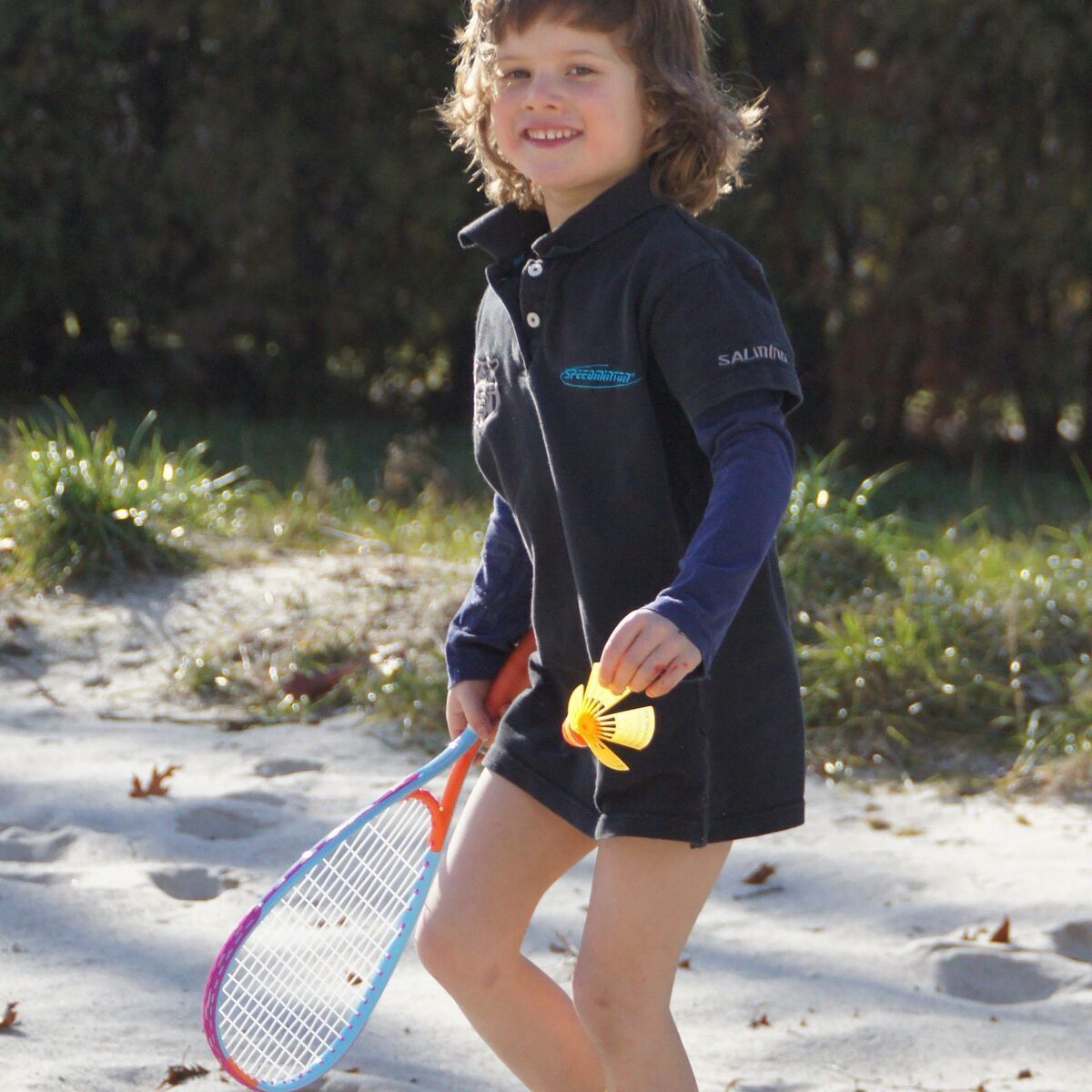Ein Kind spielt draußen im Sand mit einem Speedminton-Schläger und einem bunten Federballspiel-Set.