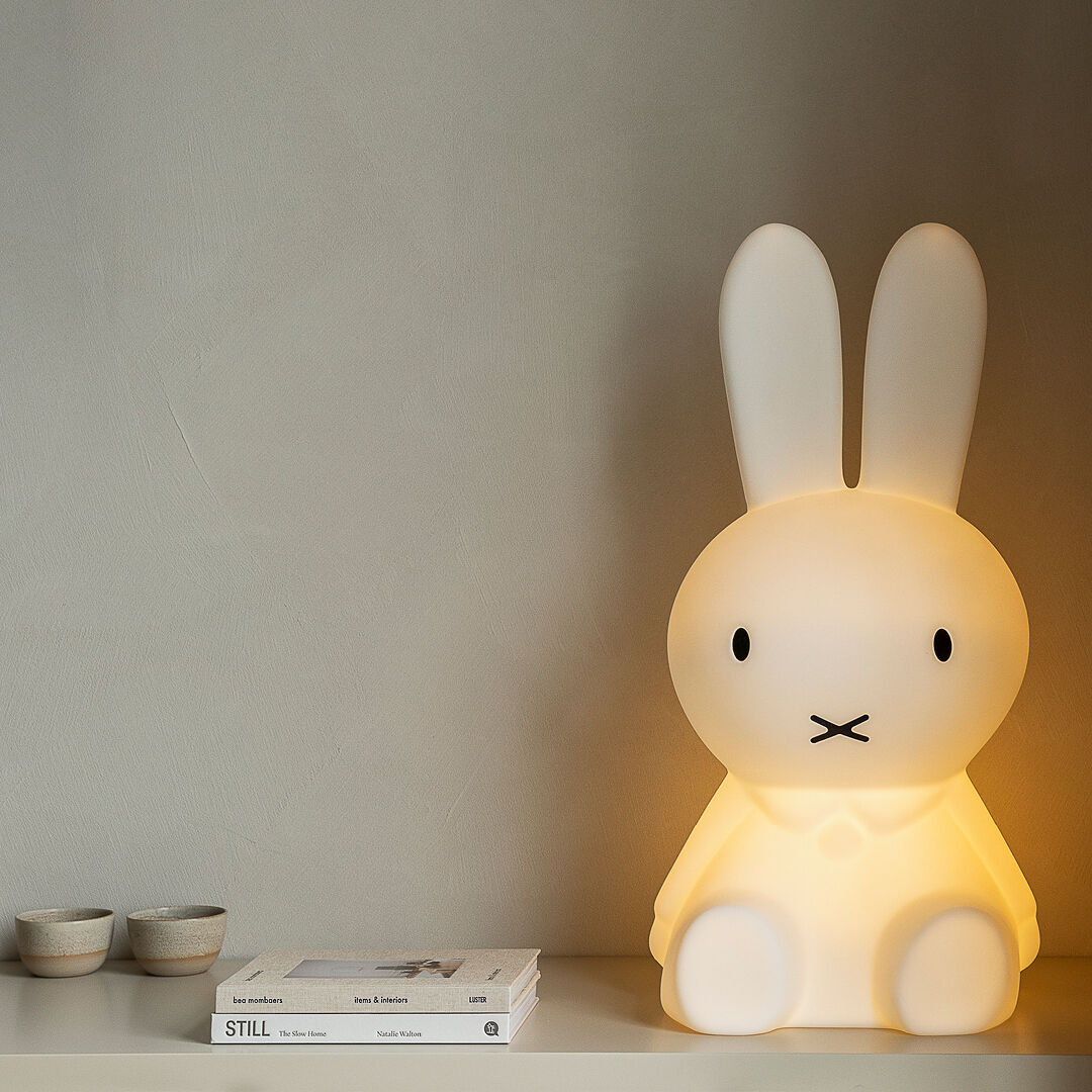 Das Bild zeigt eine »Miffy XL« Kinderlampe in Form eines großen, weißen Hasen, der auf einem Tisch neben zwei kleinen Schalen und einem Buch steht. Die Lampe leuchtet sanft und bietet eine beruhigende Beleuchtung, ideal für Kinderzimmer.