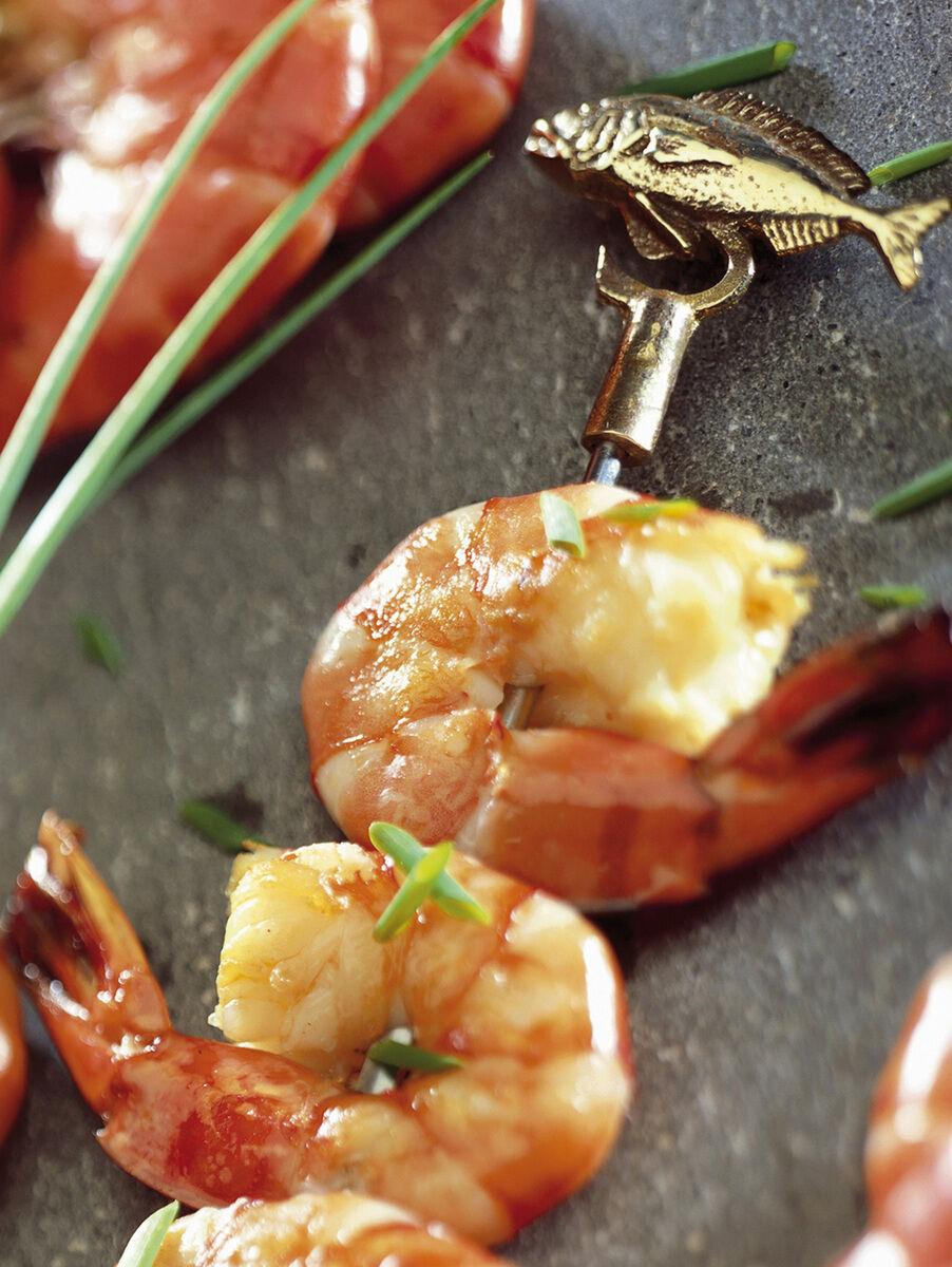 Nahaufnahme von einem Grillspieß aus Edelstahl mit gegrillten Garnelen und frischen Kräutern drauf. Im Hintergrund sind weitere Garnelen auf einem grauen Untergrund zu sehen.