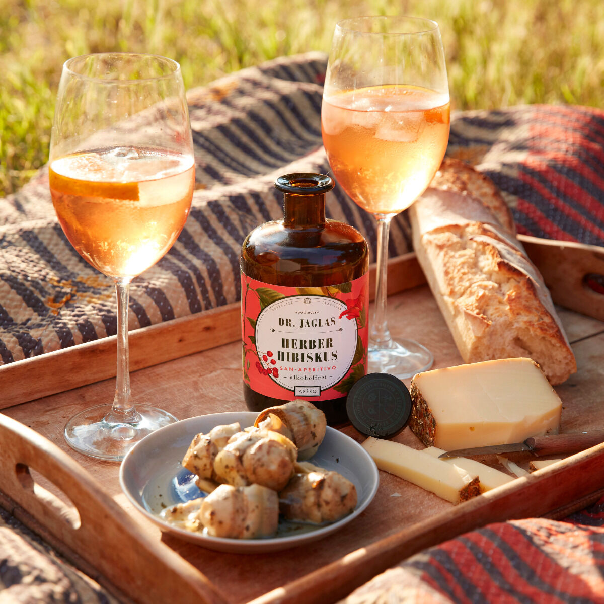 Eine Flasche »Herber Hibiskus« (alkoholfreier Aperitif) von Dr. Jaglas steht auf einem Holztablett mit zwei gefüllten Weingläsern, einer Auswahl an Brot, Käse und Kartoffeln. Im Hintergrund ist eine grüne Wiese zu sehen.