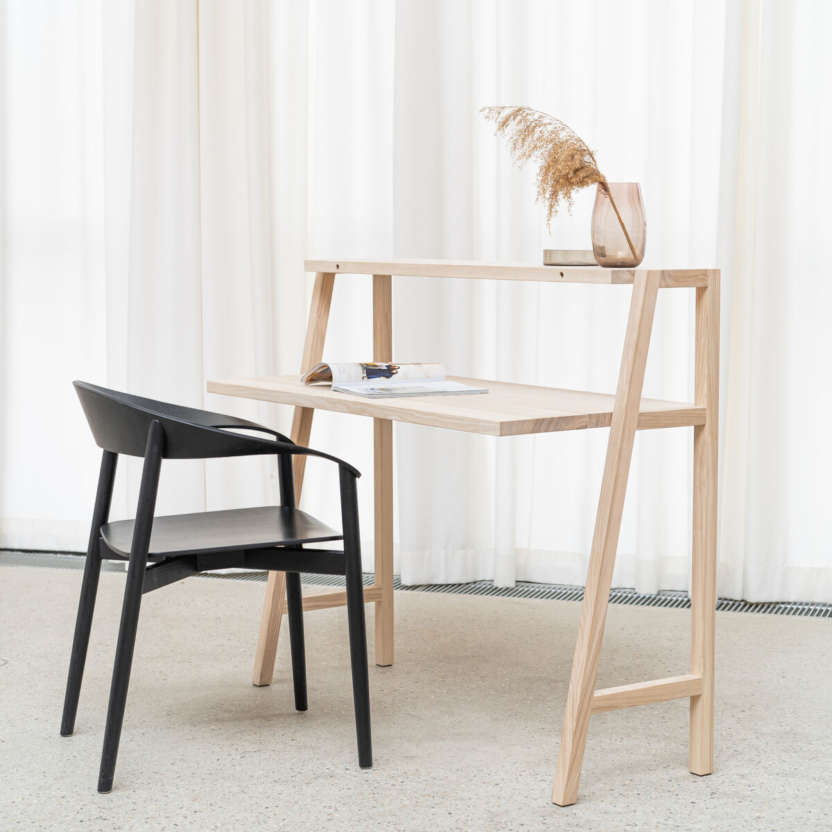 Moderner Schreibtisch KultPult "SCALA" von Frau Caze aus hellem Holz mit schrägem Gestell und Ablage, darauf eine Vase mit Trockenblume und Notizbuch, davor ein schwarzer Designstuhl, im hellen, lichtdurchfluteten Raum mit weißen Vorhängen.