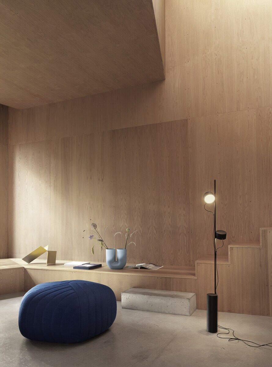 Eine moderne Bodenlampe »Post« in einem minimalistischen Innenraum mit Holzoptik. Ein runder, blauer Sitzpouf, ein weißer Betonsockel und ein hellblauer Zierkrug sind ebenfalls im Raum zu sehen. Die Lampenstruktur ist schwarz, und eine LED-Leuchte strahlt weiches Licht ab.