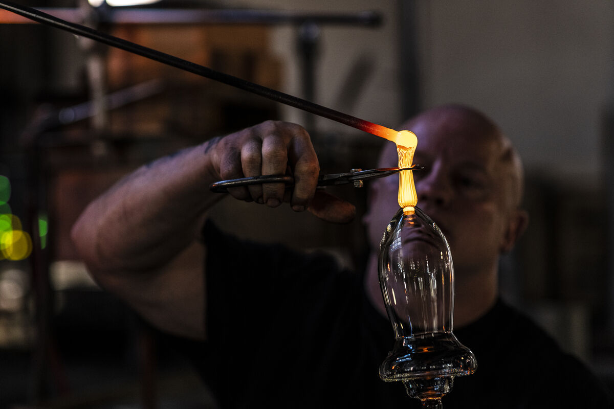 Ein Handwerker stellt mit einer heißen Flamme ein Glas her, indem er es erhitzt und formt. Daneben sind Werkzeuge und weitere Glasarbeiten zu sehen.