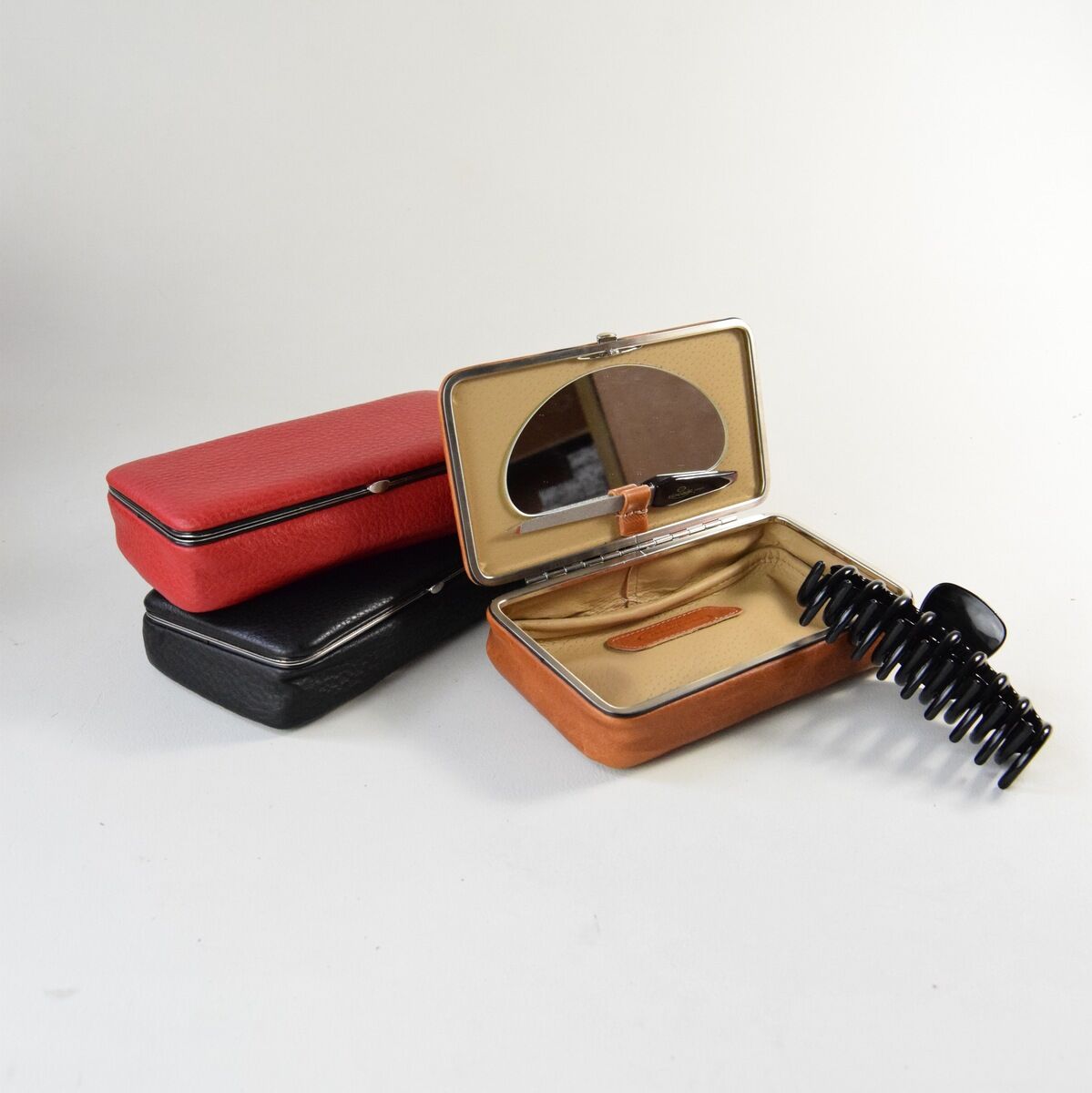 Mehrere Utensilienboxen aus Leder in unterschiedlichen Farben: eine rote und eine schwarze Box sind geschlossen, und eine hellbraune Box ist offen und zeigt ihren beigen Innenraum mit einem Spiegel im Deckel und einem Haarkamm.