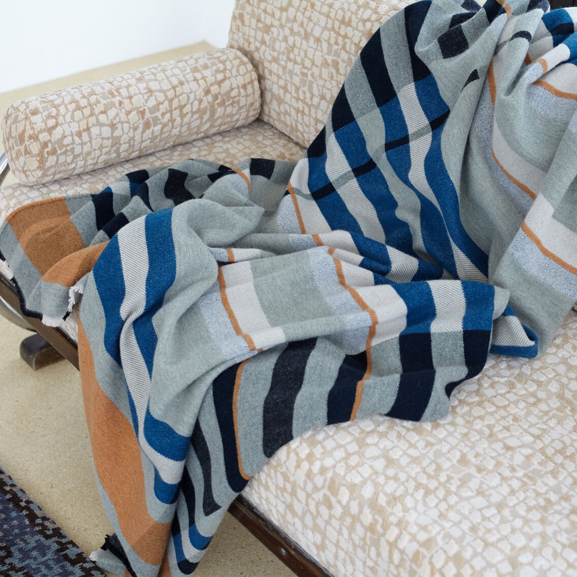 Eine gestreifte Decke in Blau-, Grau-, Weiß-, Schwarz- und Orangetönen liegt drapiert auf einem hell gemusterten Sofa. Eine weitere gemusterte Decke ist teilweise sichtbar.