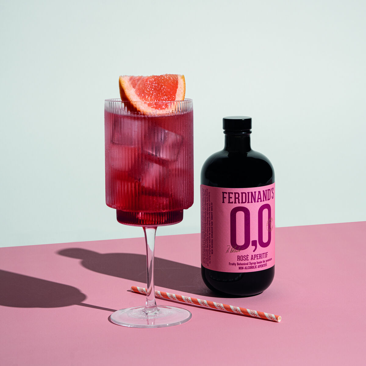 Flasche Ferdinand's 0,0 Rosé Aperitif Cordial und ein gefülltes Glas mit Eiswürfeln und einer Grapefruitscheibe auf einem rosa Tisch.