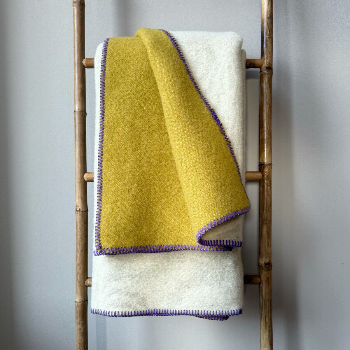 Eine Doubleface-Decke mit einer gelben und weißen Seite, dekoriert mit lila Rändern, hängt über einer Bambusleiter vor einer grauen Wand.