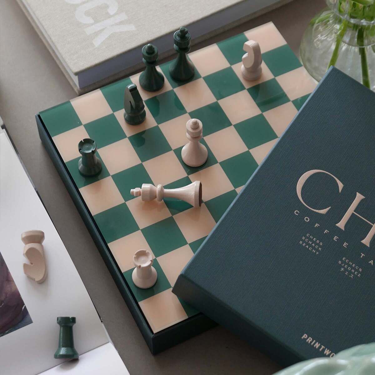 Auf einem Tisch liegt ein klassisches Schachspiel mit grün-beigem Schachbrett und passenden Schachfiguren. Das Spielfeld ist teilweise von einer stilvollen Verpackung mit der Aufschrift 'CH' verdeckt. Umschlossen wird das Schachspiel von Büchern und einer Vase, die eine gemütliche und einladende Atmosphäre betonen.