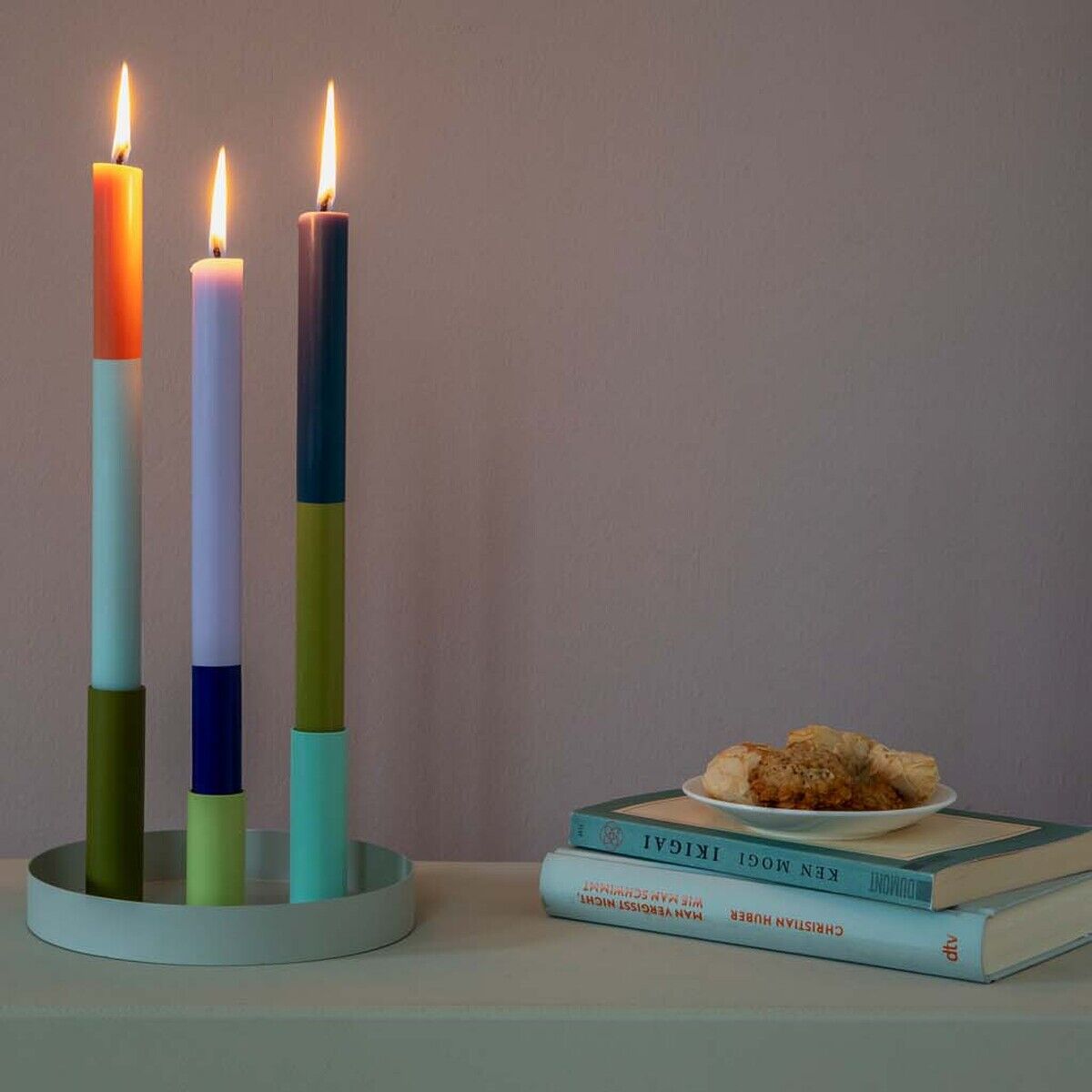 Ein Set von drei brennenden Stabkerzen in unterschiedlichen Farben (Orange, Blau, Lila, Grün, Weiß) auf einem runden Kerzenhalter auf einem Tisch. Daneben liegen einige Bücher und ein kleiner Teller mit Gebäck.