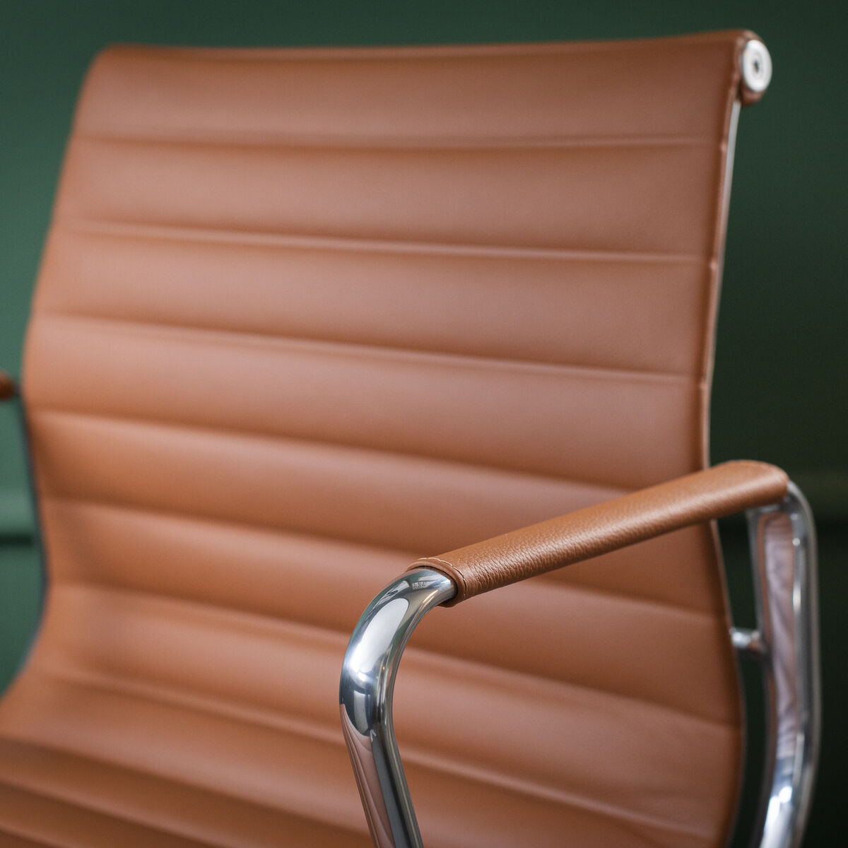 Nahaufnahme eines Vitra Alu Chairs EA 118. Der Bürostuhl hat eine braune Lederpolsterung und polierte Armlehnen aus Metall. Der Hintergrund ist grün.