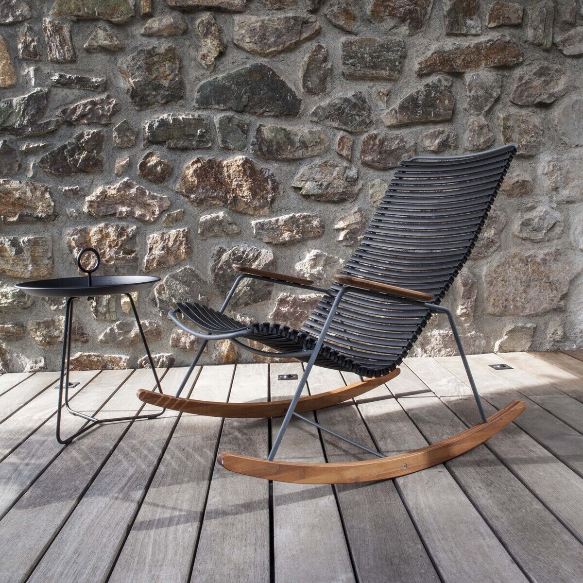 Ein stilvoller Tray Table »EYELET« Beistelltisch aus schwarzem Metall steht auf einer Terrasse mit Holzboden vor einer rustikalen Steinwand. Daneben befindet sich ein moderner Schaukelstuhl mit schwarzen und braunen Akzenten.