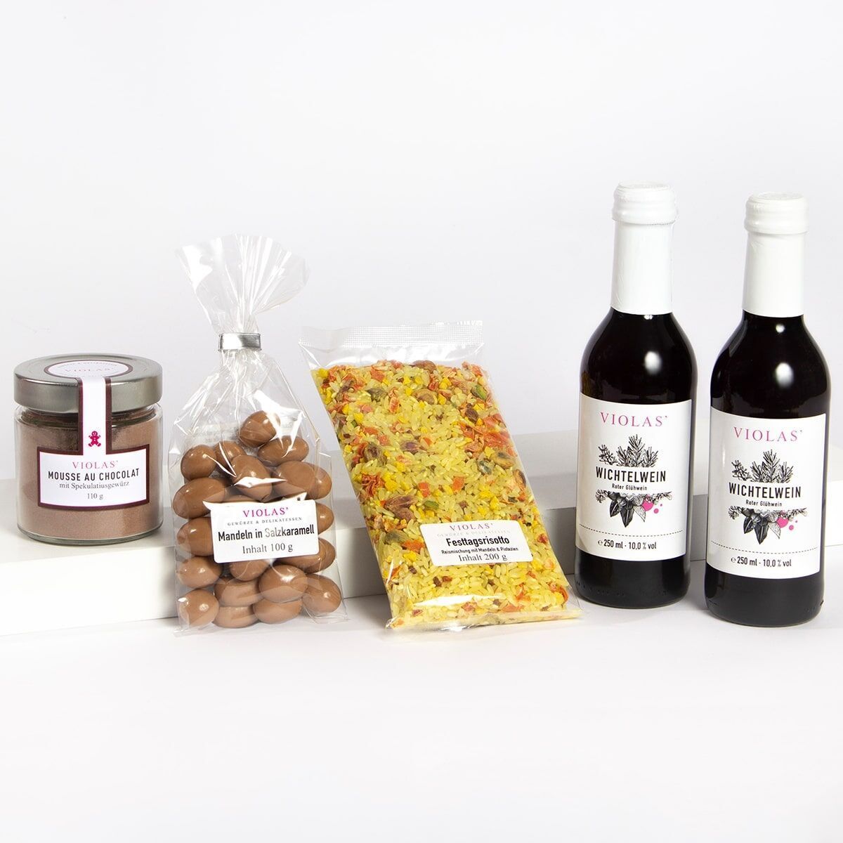 Weihnachtszeit-Set bestehend aus einem Glas Mousse au Chocolat, einer Tüte mandeln in Vollmilchschokolade, einer Packung Festtagspasta und zwei Flaschen Wichtelweinen. Alle Produkte werden auf einem weißen Untergrund arrangiert dargestellt.