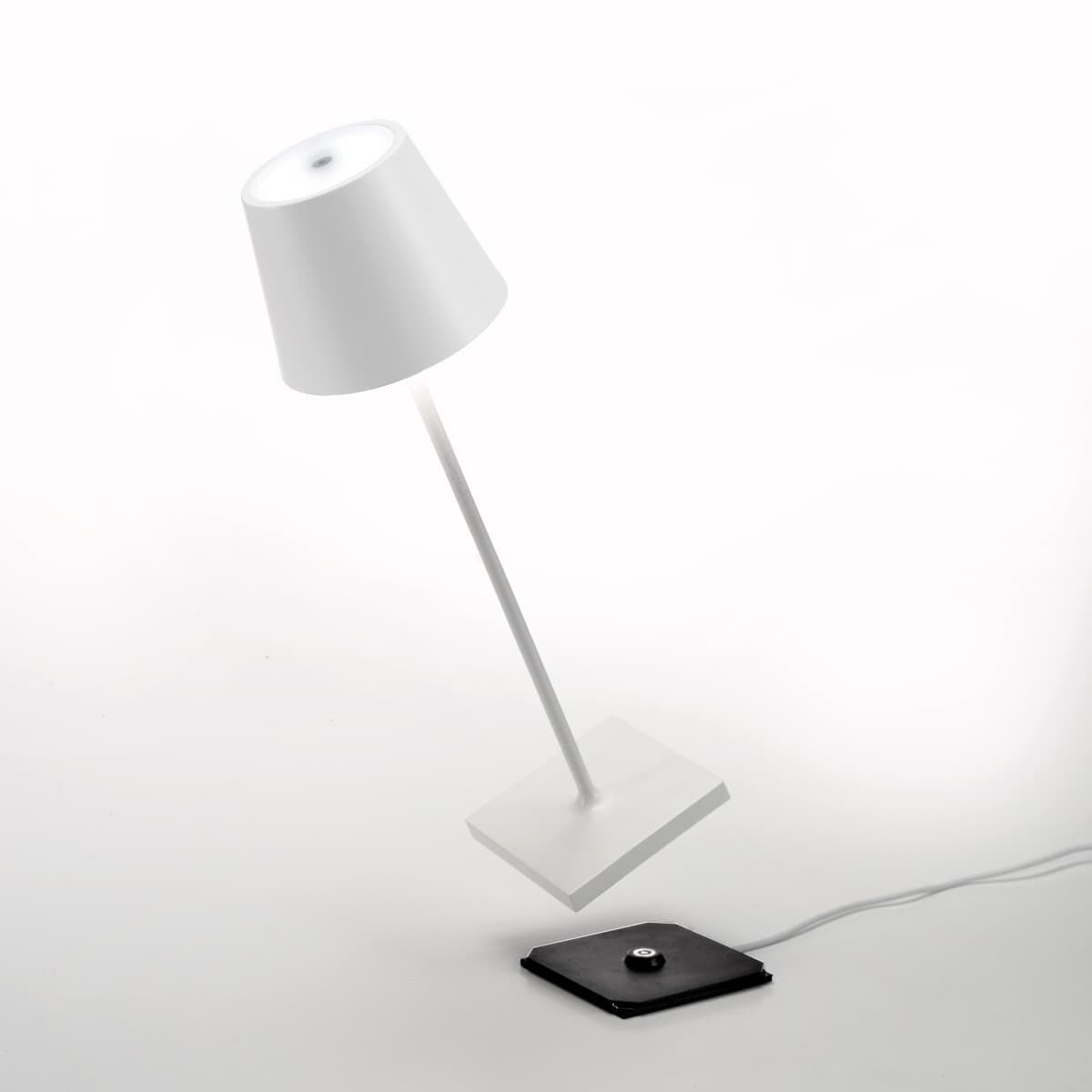 Eine weiße LED-Tischlampe »Poldina Pro« mit einem schlanken, minimalistischen Design, die auf einer quadratischen Basis steht und über ein schwarzes USB-Ladekabel verfügt.