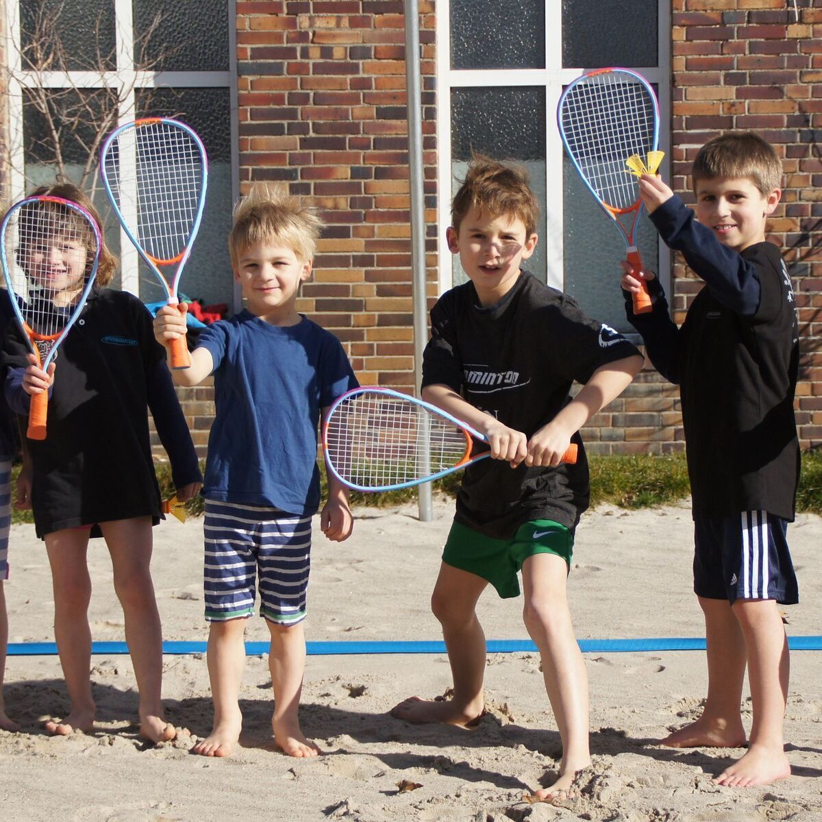 Fünf Kinder stehen auf einem sandigen Untergrund vor einem Backsteingebäude und halten bunte Speedminton-Schläger. Sie tragen sportliche Kleidung und lächeln in die Kamera. Das Wetter ist sonnig, und alle sehen glücklich aus.
