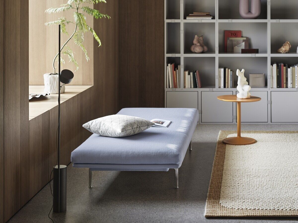 Modernes Wohnzimmer mit einer Bodenlampe 'Post'. Hellblaue Chaiselongue mit Kissen, daneben ein Holzbeistelltisch mit Deko. Im Hintergrund ein Regal mit Büchern und Dekorationsobjekten.