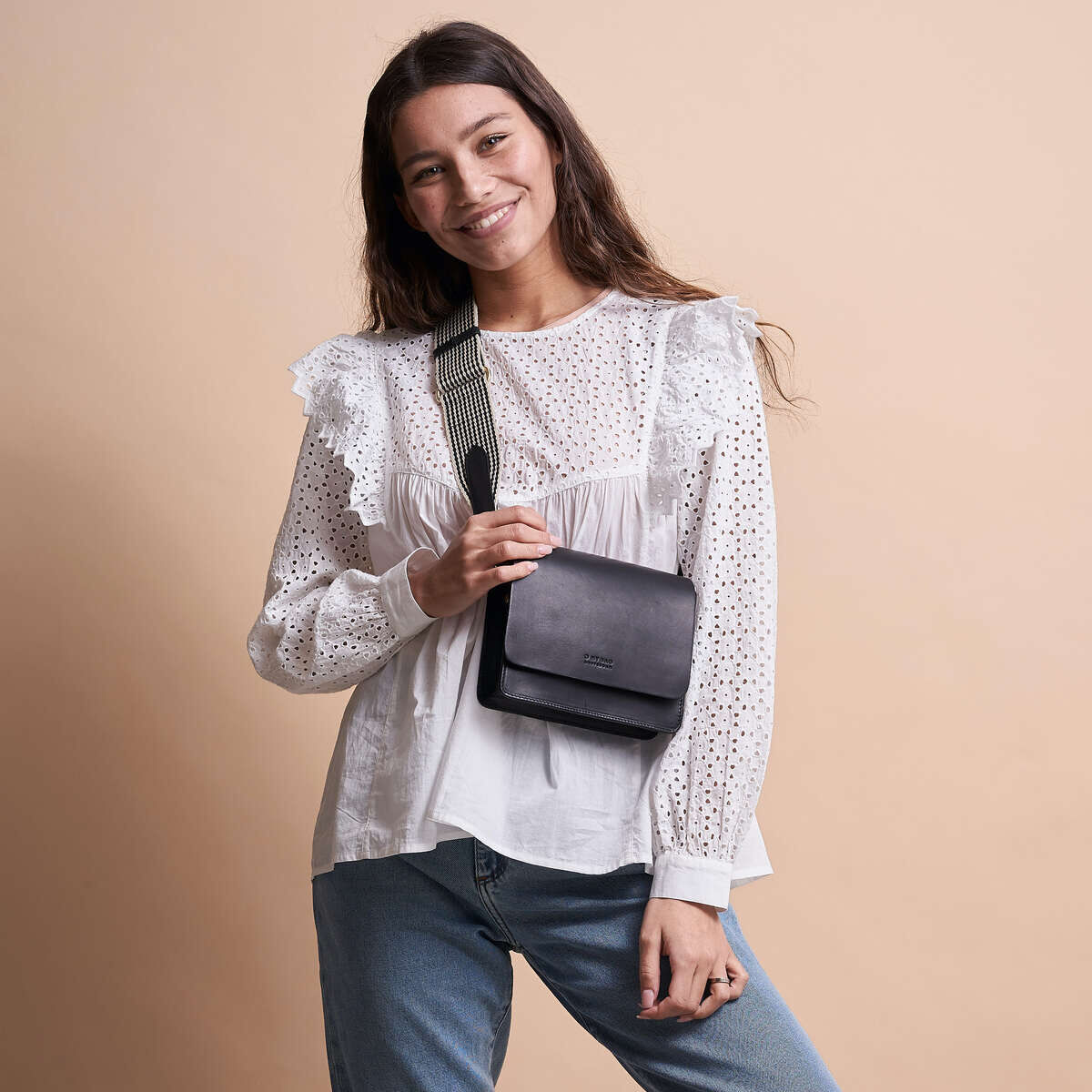 Junge Frau trägt eine weiße, spitzenbesetzte Bluse und blauen Jeans und hält eine schwarze Tasche »Audrey Mini« mit Crossbody-Riemen, vor einem beigefarbenen Hintergrund.