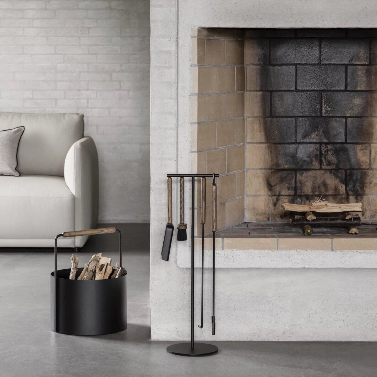 Moderner Holzkorb aus schwarzem Metall mit Holzhenkel steht neben einem offenen Kamin in einem minimalistisch eingerichteten Wohnzimmer mit heller Couch und grauen Wänden. Der Korb ist mit Feuerholz gefüllt und vermittelt ein wohnliches, skandinavisches Ambiente.