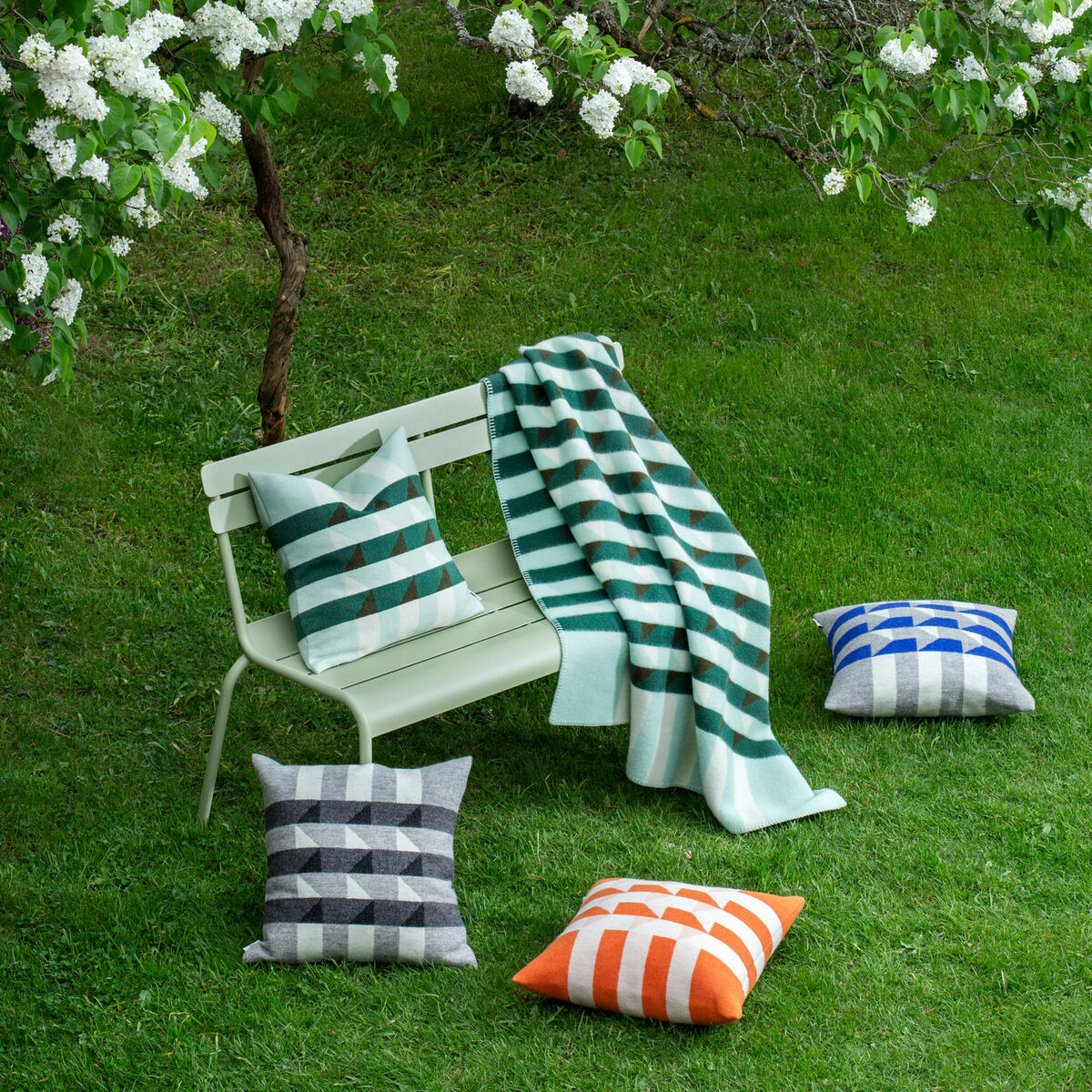 Ein bequemer, weißer Gartenstuhl unter einem blühenden Baum. Auf dem Stuhl befindet sich ein gestreiftes Kissen und eine passende Decke. Weitere gestreifte Kissen in Blau, Orange, Grau und Grün liegen auf dem Gras daneben. Perfekt für eine Sommer- oder Gartenparty.
