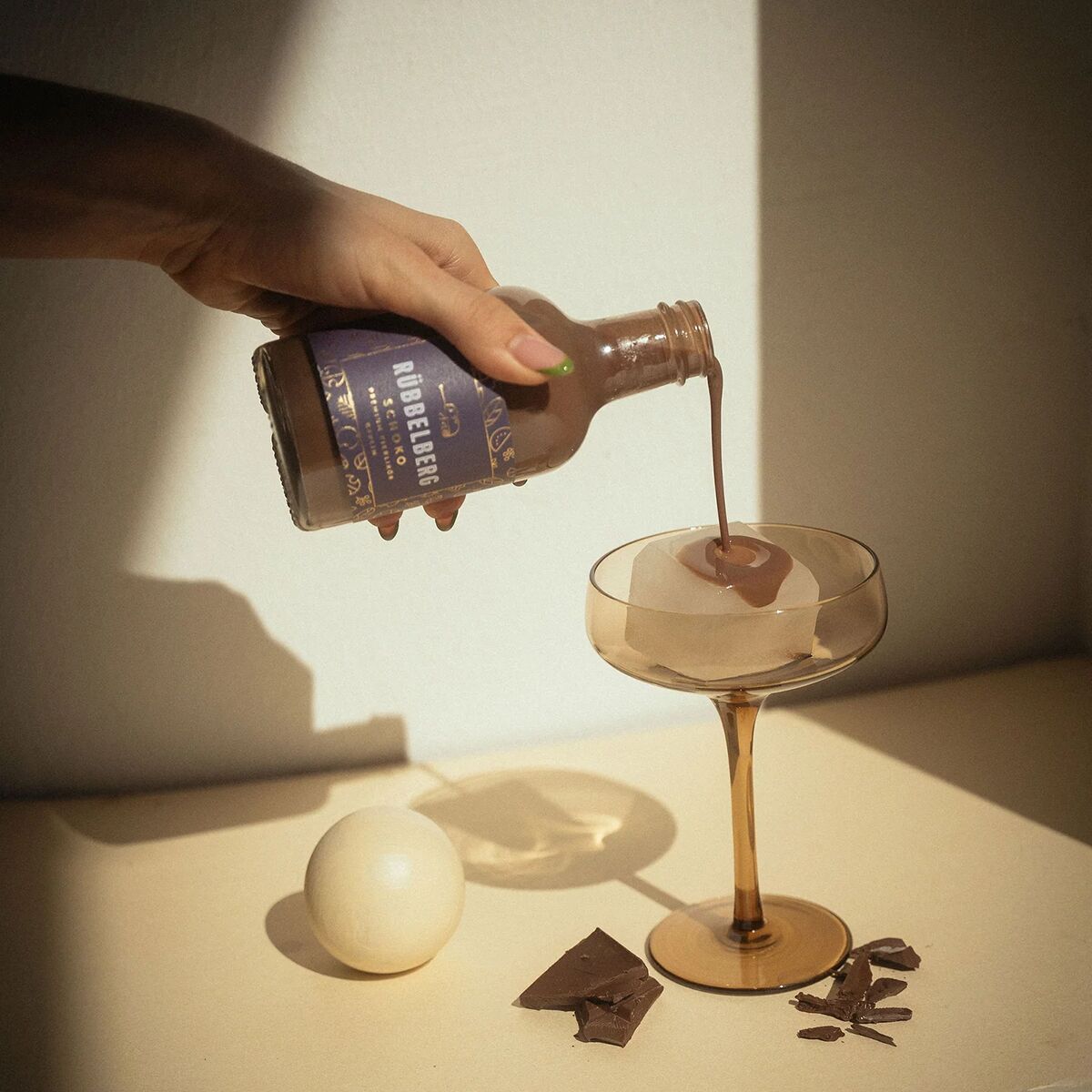 Eine Hand gießt Premium Eierlikör Schoko aus einer braunen Flasche in ein elegantes Glas. Neben dem Glas liegen Stücke dunkler Schokolade und ein rundes, weißes Objekt auf einer beige Unterlage. Die Szene ist warm beleuchtet und vermittelt eine festliche Stimmung.