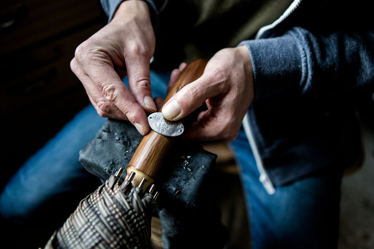 Nahaufnahme von Händen, die ein Metallschild an einem handgefertigten Holzgriff eines Regenschirms befestigen. Der Schirmgriff zeigt feine Handwerkskunst und Detailarbeit.