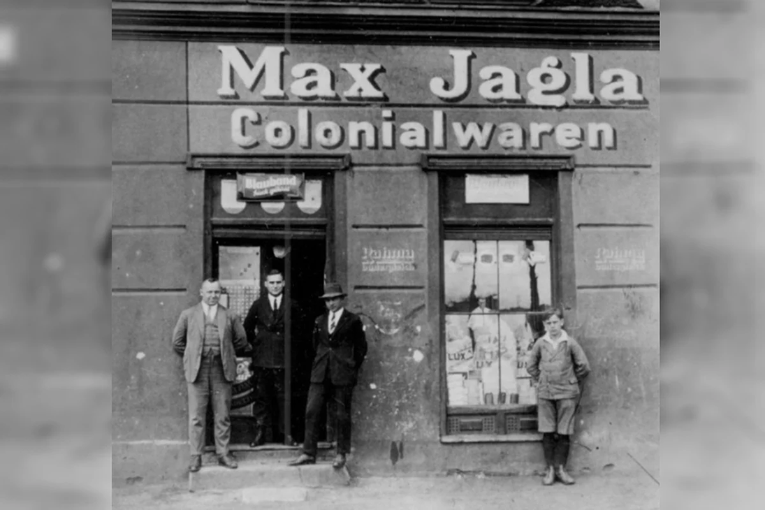 Vier Personen vor einem Geschäft mit der Aufschrift 'Max Jagla Colonialwaren', drei Männer und ein Junge, historische Schwarzweißaufnahme