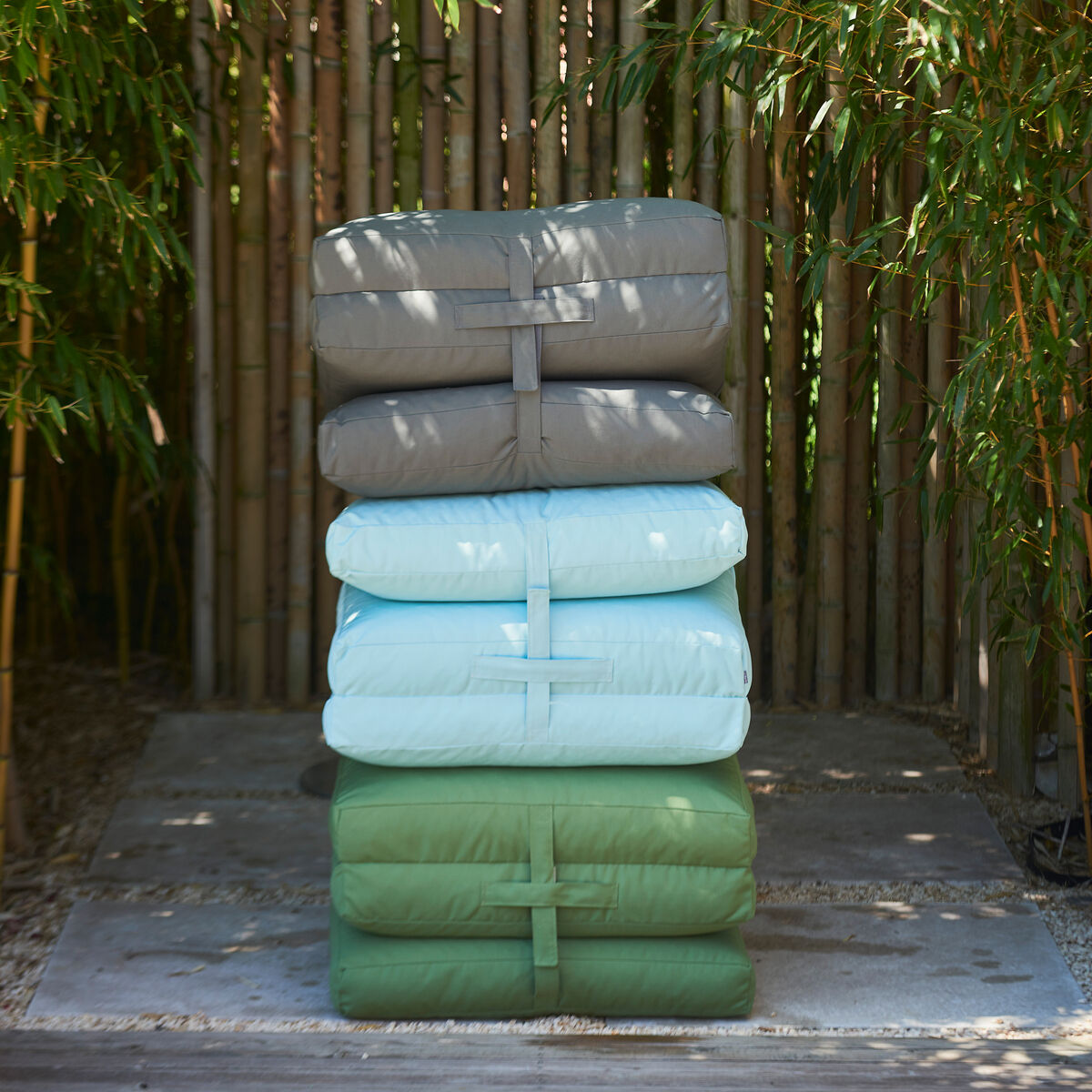 Eine gestapelte Gruppe von drei Gartenmatratzen in den Farben Grau, Blau und Grün steht in einem Außenbereich vor einem Hintergrund aus Bambus.