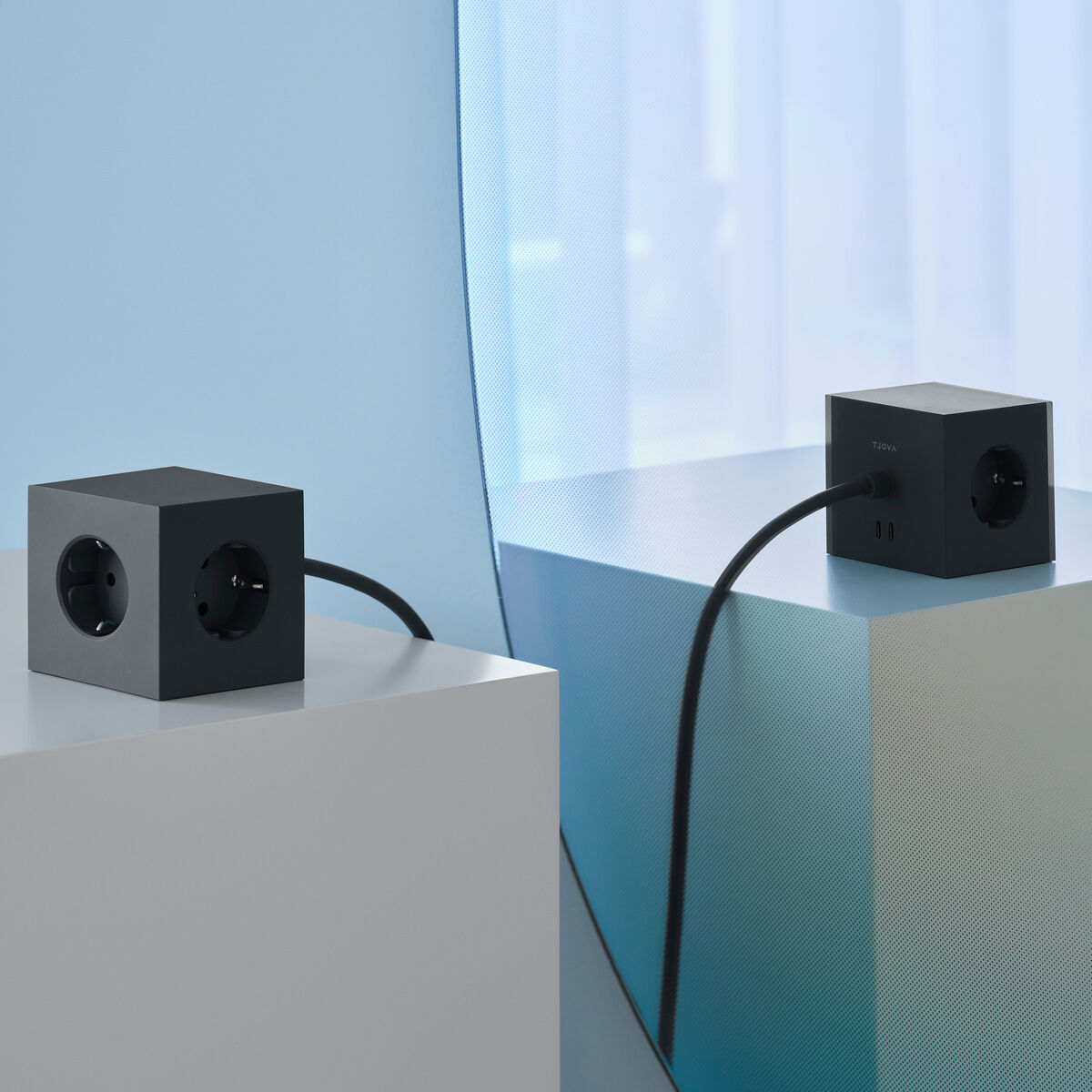 Das Bild zeigt zwei schwarze quadratische Geräte auf weißen Podesten. Diese Geräte haben USB-C-Anschlüsse und Magneten, was auf vielseitige Anschlussmöglichkeiten hinweist. Der Hintergrund ist blau-weiß und vermittelt einen modernen und minimalistischen Eindruck, der perfekt für Büro- oder Konferenzraumeinrichtungen geeignet ist.
