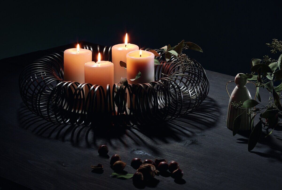 Ein moderner Adventskranz namens »Caracas« aus schwarzem Metall, der vier weiße Kerzen hält, die brennen. Der Kranz ist auf einem dunklen Holztisch platziert, umgeben von einigen Tannenzweigen, Nüssen und einem kleinen Engel aus Holz. Die Szene verströmt eine gemütliche und feierliche Stimmung.