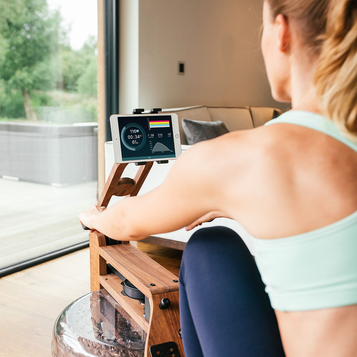 Eine Frau trainiert auf einem Waterrower-Rudergerät aus Eiche, während sie in einem modernen Wohnzimmer sitzt. Sie betrachtet ein digitales Display, das auf dem Gerät angebracht ist. Im Hintergrund ist eine große Fensterfront mit Blick nach draußen sichtbar.