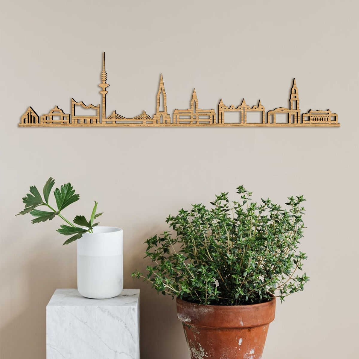 Eine minimalistische Wanddekoration aus Holz zeigt die Skyline von Hamburg. Die Dekoration hängt an der Wand über zwei Pflanzen in unterschiedlichen Höhen.