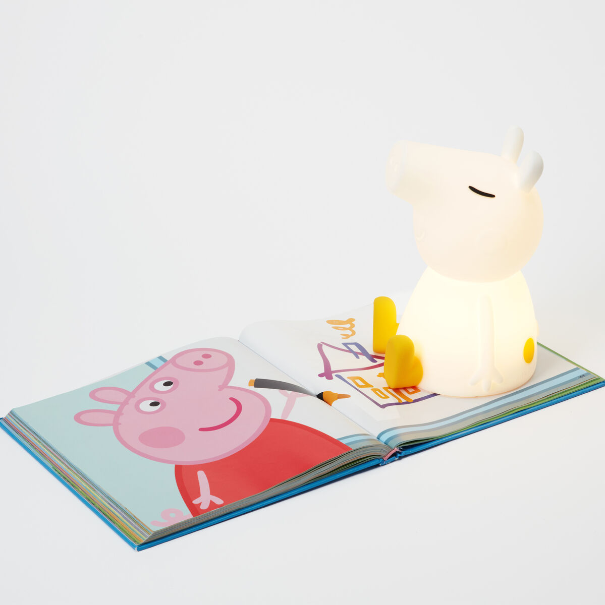 Ein Nachtlicht in Form einer Figur von Peppa Pig, das neben einem aufgeschlagenen Buch steht. Die Figur leuchtet sanft und erzeugt eine beruhigende Atmosphäre. Die Farben des Buches sind überwiegend hell mit Blau und Rosa.