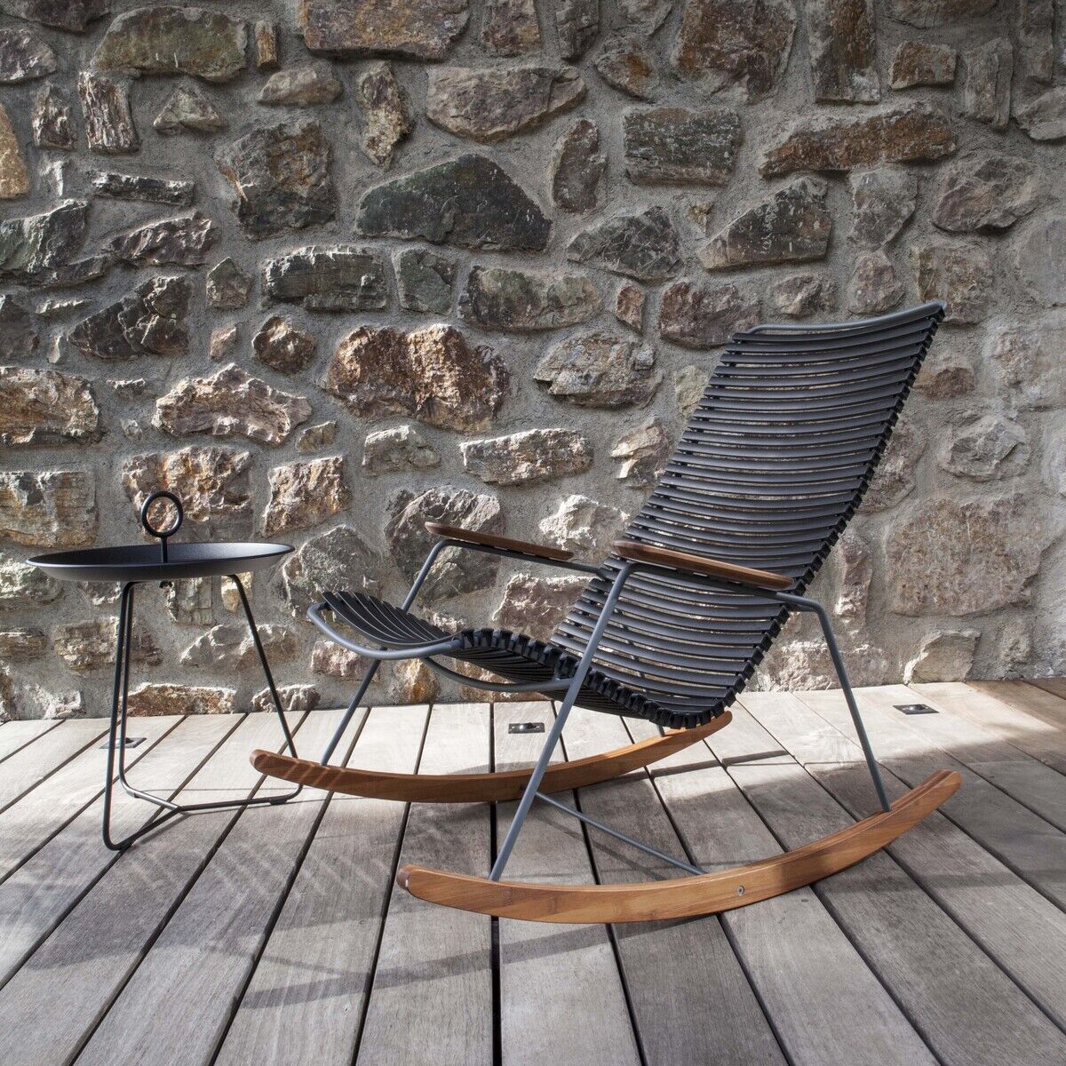 Schaukelstuhl mit schwarzem Metallgestell und Holzelementen auf einer Holzterrasse neben einem kleinen Beistelltisch und einer Steinwand im Hintergrund.