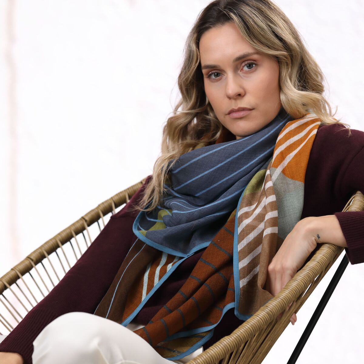 Frau sitzt entspannt in einem modernen Sessel und trägt einen mehrfarbigen Wollschal mit blauen, braunen, orangen und grünen Streifen. Der Schal liegt elegant um ihre Schultern und betont ihr herbstliches Outfit, bestehend aus weinrotem Oberteil und heller Hose.