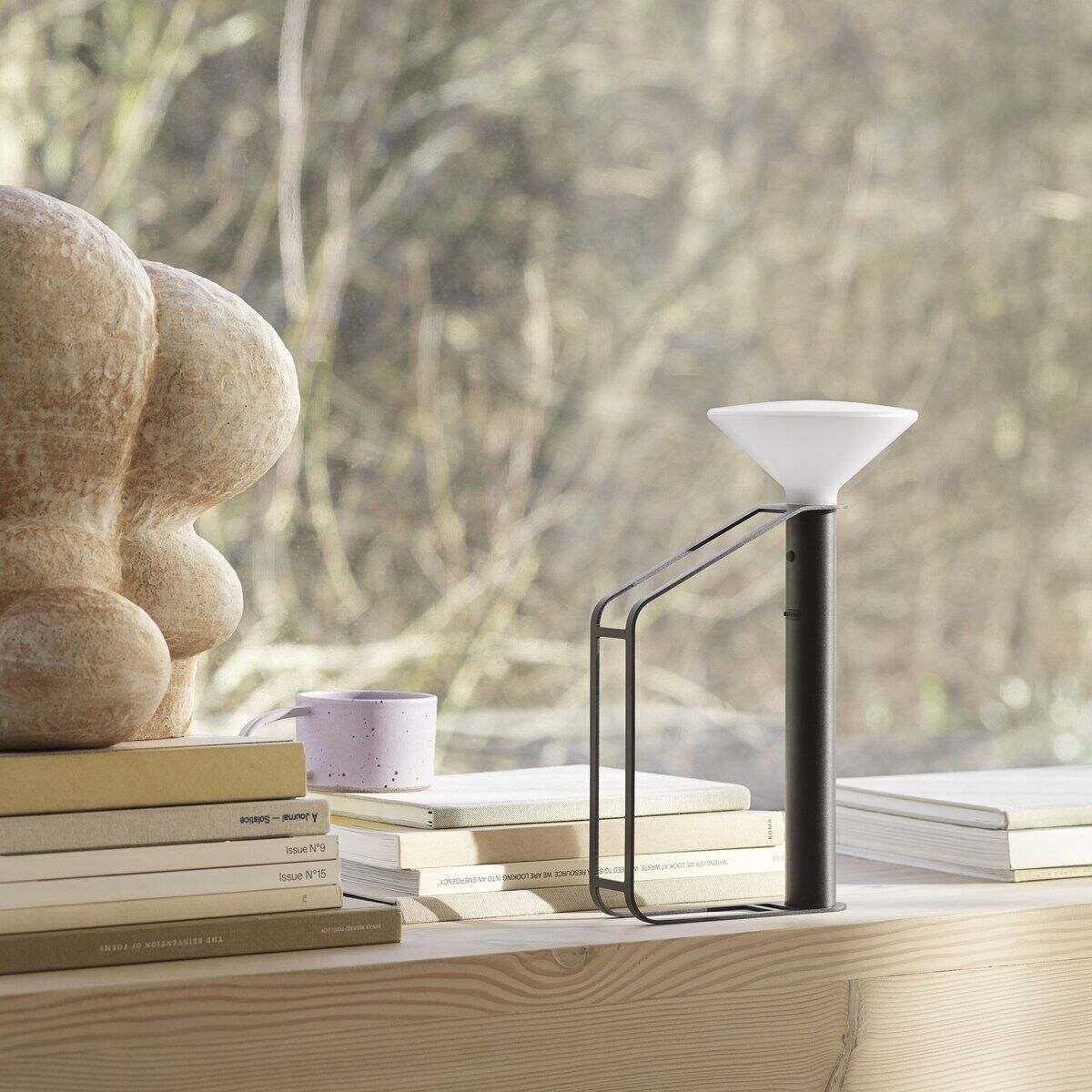 Eine tragbare Lampe namens 'Piton' steht auf einem Tisch neben einem Stapel Bücher und einer Skulptur. Die Lampe hat ein modernes und minimalistisches Design mit einem schwarzen Ständer und einem weißen Lampenschirm. Im Hintergrund sind Bäume zu sehen, die durch ein großes Fenster hindurch sichtbar sind.