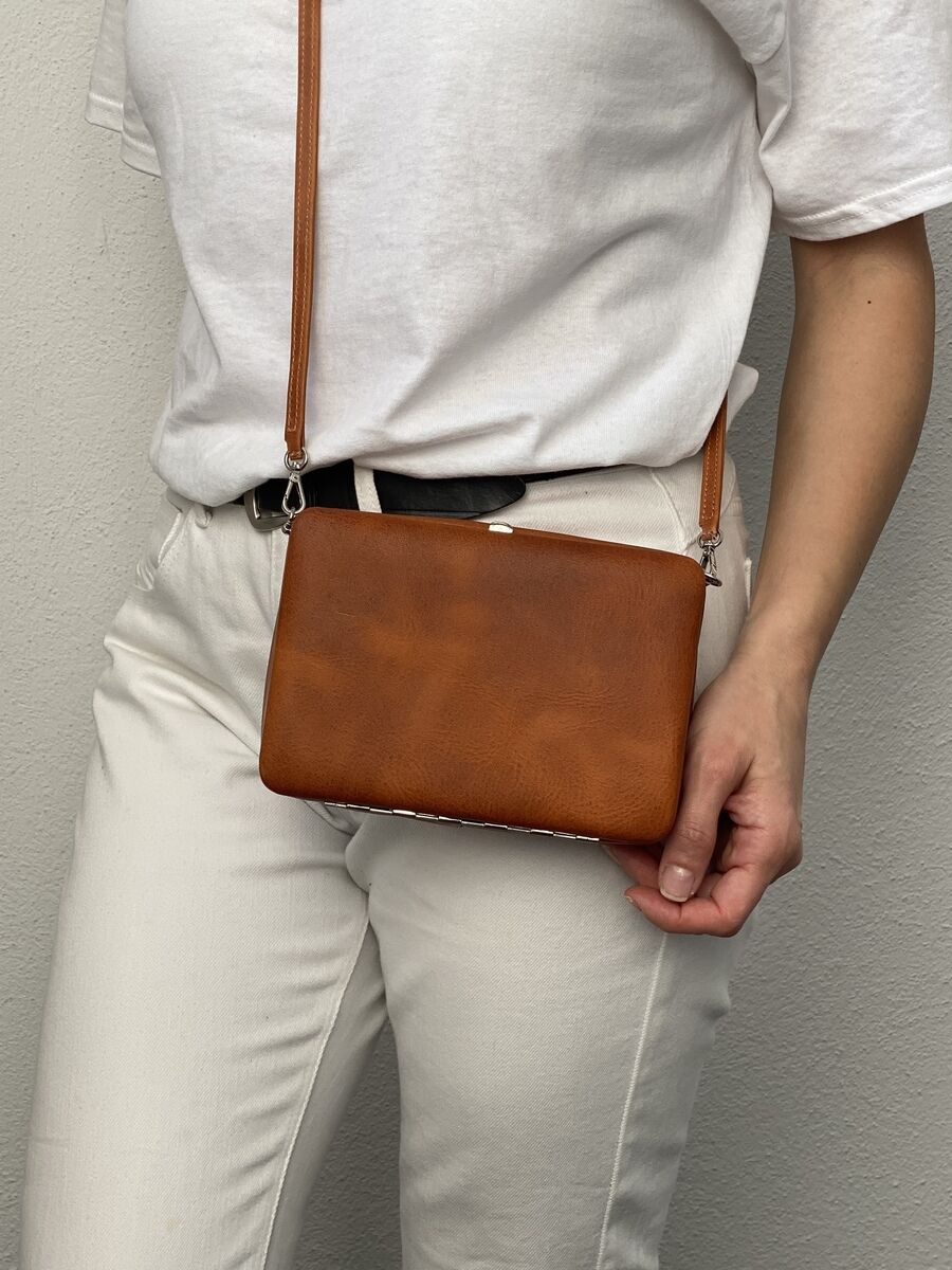Frau mit einer braunen Leder-Umhängetasche namens ZEIT-Tasche »Jacky«, die quer über den Körper getragen wird. Die Frau trägt ein weißes T-Shirt und eine weiße Hose.