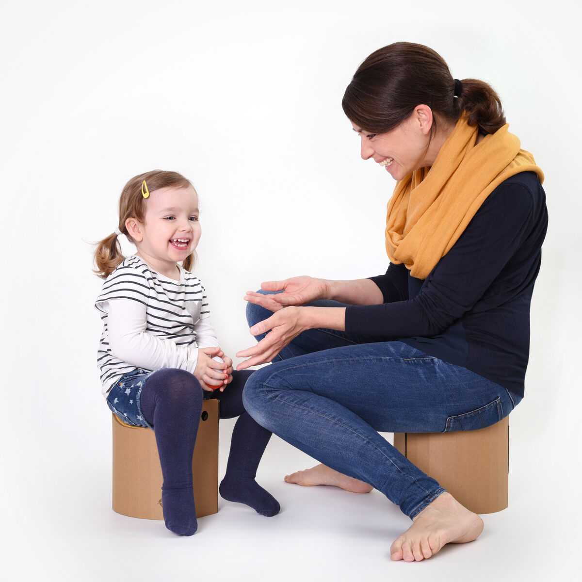 Eine Mutter und ihr kleines Kind sitzen auf Papphockern in einem minimalistischen Innenraum. Die Frau trägt ein dunkelblaues Langarmshirt und einen gelben Schal, während das Kind eine gestreifte Bluse und marineblaue Leggings anhat. Beide wirken glücklich und genießen den Moment miteinander.