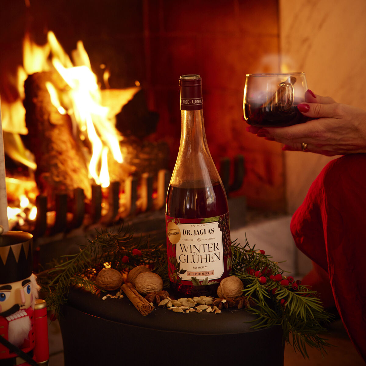 Im Vordergrund steht eine Flasche Dr. Jaglas Winterglühen Merlot 3er Winzerglühwein alkoholfrei auf einem Tisch, umgeben von Tannenzweigen, Zimtstangen, Nüssen und weihnachtlicher Dekoration. Dahinter brennt ein Kaminfeuer, eine Hand hält ein mit Glühwein gefülltes Glas.