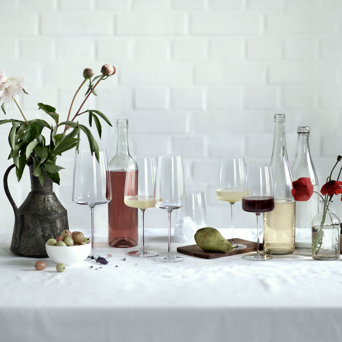 Attraktive Tischdekoration mit eleganten Weingläsern aus Kristall, gefüllt mit Sekt und Wein. Im Hintergrund befinden sich Weinflaschen und eine Vase mit grünen Pflanzen und roten sowie rosa Blumen. Auf dem Tisch liegen verschiedene Früchte und Beeren. Eine friedliche und einladende Atmosphäre, perfekt für Feierlichkeiten und besondere Anlässe.