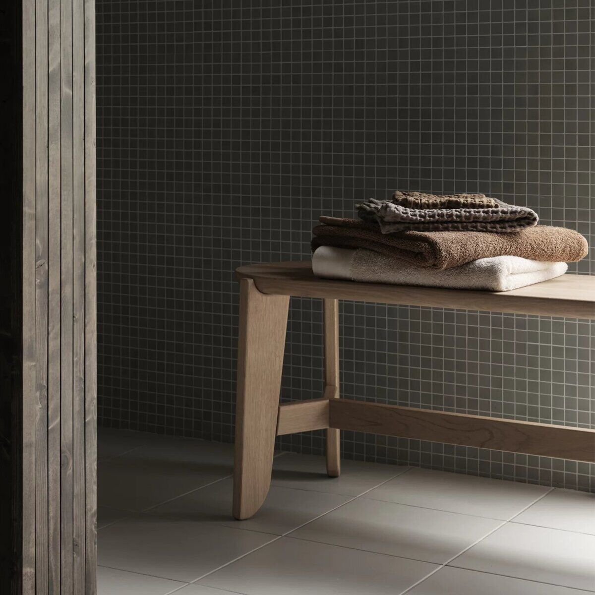Helle Holzbank im minimalistischen Badezimmer vor gefliestem Hintergrund, auf der mehrere ordentlich gefaltete Handtücher in neutralen Farben liegen. Die Gesamtstimmung ist ruhig und entspannend, passend für moderne, skandinavische Wohnstile.