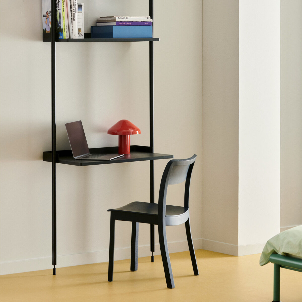 Moderne, tragbare Lampe in Rot auf einem schwarzen Schreibtisch mit einem Laptop, zusammen mit einem schwarzen Stuhl. Hintergrund sind weiße Wände und offene Regale mit Büchern.