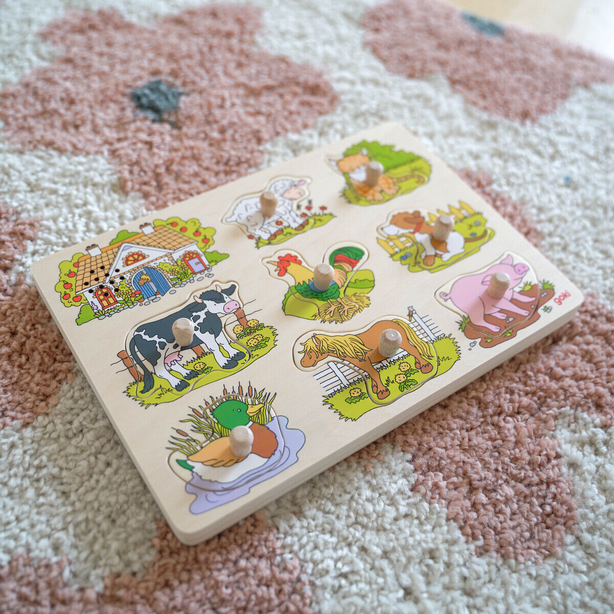 Abgebildet ist ein Holzpuzzle mit Bauernhoftieren. Die Puzzleplatte zeigt bunte Illustrationen von Tieren wie Kuh, Schwein, Schaf, Hahn, Ente und Pferd. Jedes Puzzleteil ist mit einem Knopf zum Greifen versehen. Der Hintergrund besteht aus einem hellen Teppich mit rosa und weißen Mustern.