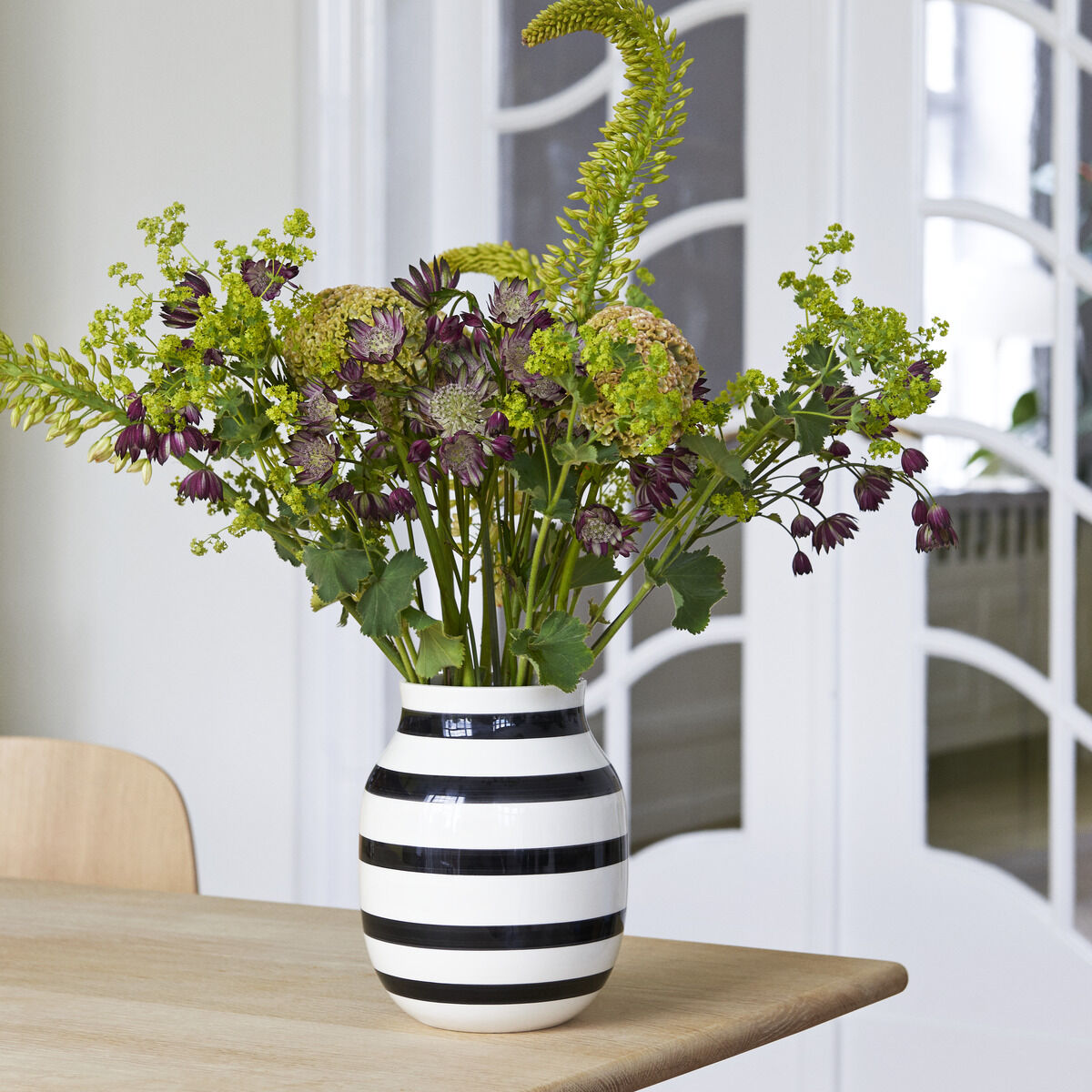 Schwarz-weiß gestreifte Vase »Omaggio H20« auf einem Tisch, gefüllt mit einem bunten Blumenarrangement aus grünen, violetten und gelben Blüten. Im Hintergrund sind weiße Türen mit Glasscheiben zu sehen.