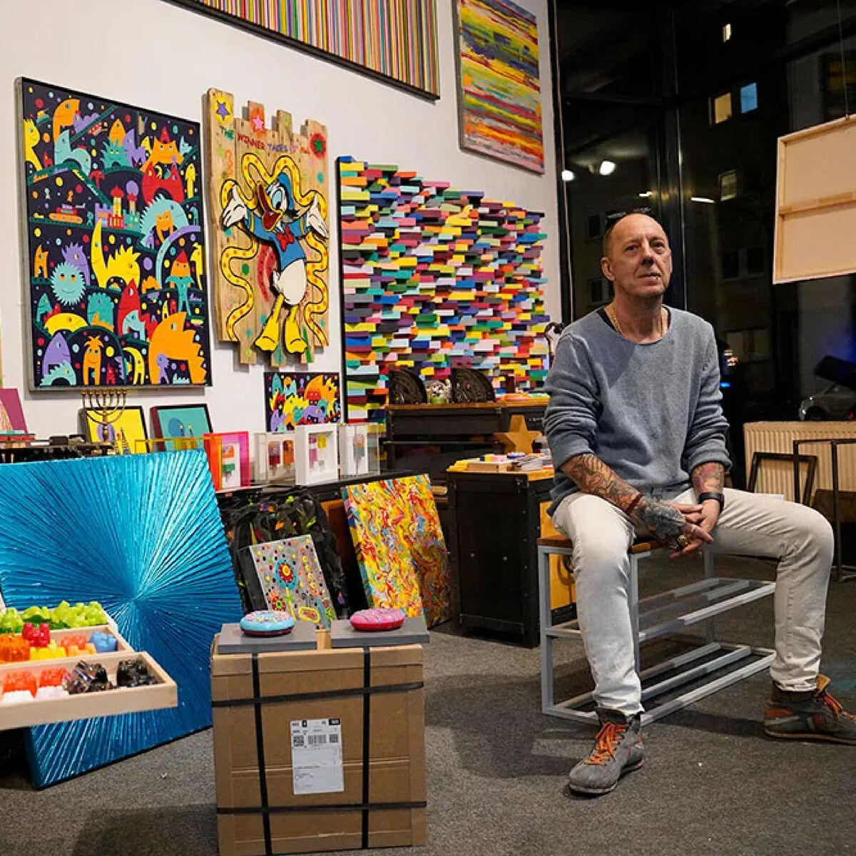 Ein Künstler sitzt in seinem Atelier, umgeben von farbenfrohen Pop-Art-Gemälden und plastischen Arbeiten, die Comics, abstrakte Grafiken und leuchtende Farben zeigen. Werke sind an den Wänden und auf Möbeln arrangiert, viele Details und kreative Inspirationen füllen den Raum.