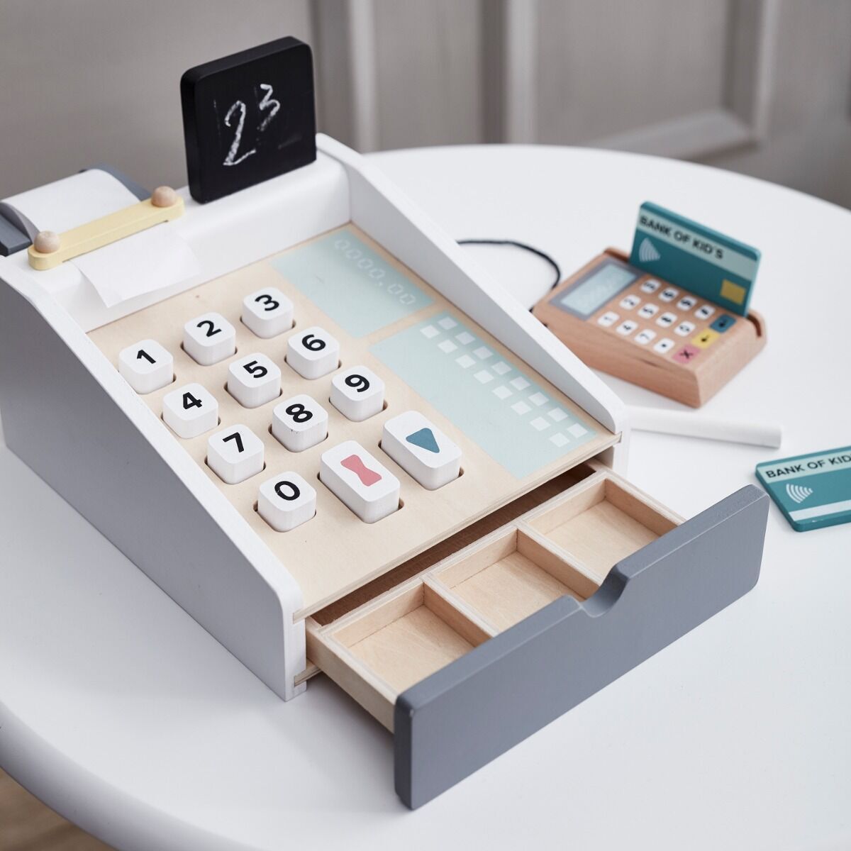 Holzkasse mit weißen und blauen Tasten, einer kleinen Tafel und Schubladen, die auf einem weißen Tisch steht. Im Hintergrund befinden sich weiteres Spielzubehör wie eine Spielkarte und ein Kartenlesegerät.