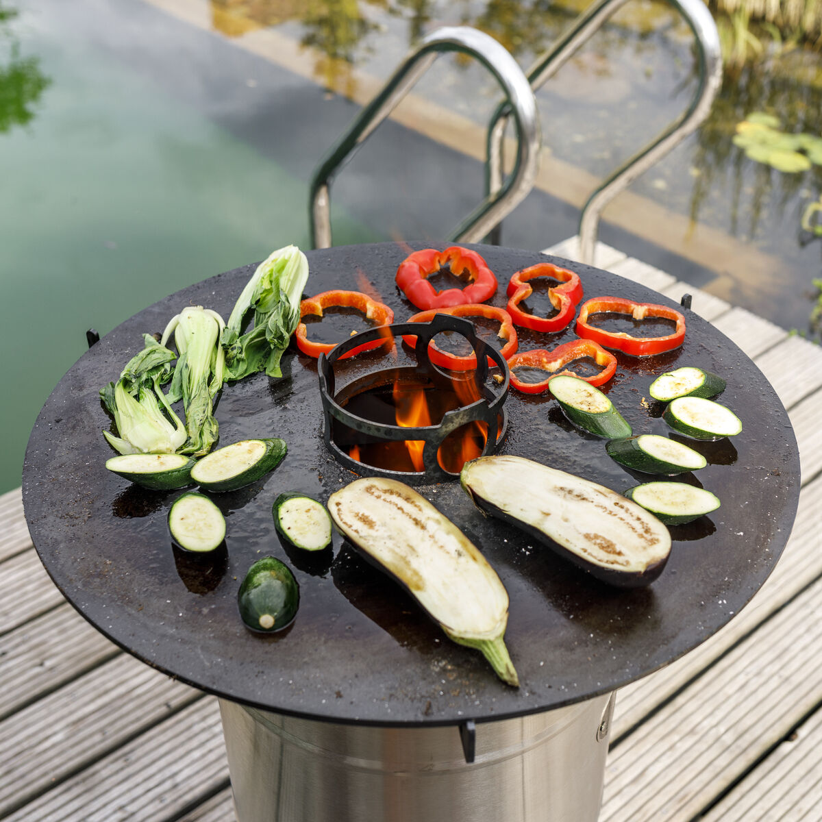 Grillplatte namens »Pyron« mit verschiedenen Gemüsesorten wie Paprika, Aubergine und Zucchini darauf. Sie steht auf einem Edelstahlgestell neben einem Teich und ist von Natur umgeben. Perfekt für Grillabende und Outdoor-Kochen.